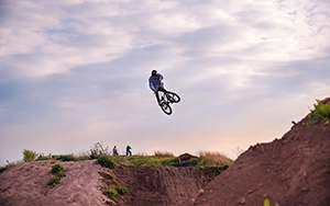Ein BMX-Fahrer mit Helm schwebt hoch in der Luft über einer Dirt-Jump-Anlage, sein Fahrrad unter ihm. Der Himmel ist bewölkt und im Hintergrund sind weitere Erdhügel und kleine Silhouetten zu sehen.